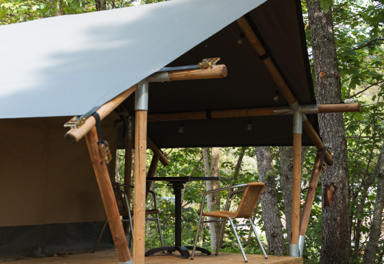 Tenda safari con veranda in legno e due sedie, immersa tra gli alberi in un ambiente boscoso verde.