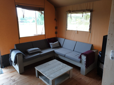 Innenansicht eines Safarizeltes mit grauem Ecksofa, Couchtisch und Fenstern auf Camping Gorishoek, Niederlande.