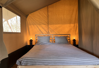 Interior of a safari tent at Camping 'de Val' with double bed, striped bedding, and warm lighting.