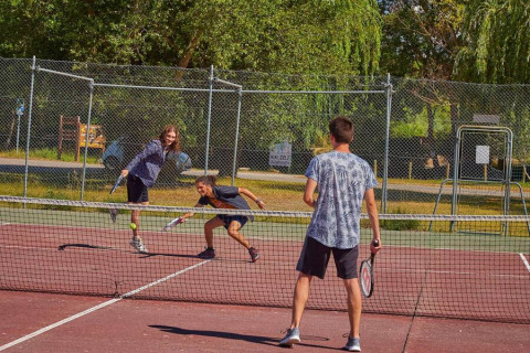 Tre personer spiller tennis på en udendørs bane omgivet af grønne træer ved Seasonova Camping Les Prés du Verdon.