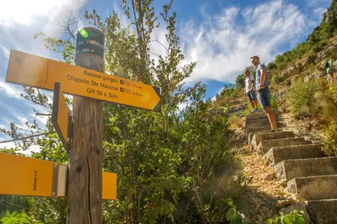 Wandelaars beklimmen stenen trappen bij Quinson, Provence, langs gele borden en groene natuur.