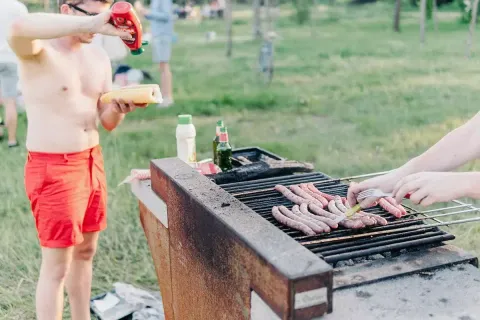 Twee mensen barbecueën worsten op een buitenbarbecue terwijl een man ketchup op zijn broodje doet.
