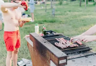 Twee mensen barbecueën worsten op een buitenbarbecue terwijl een man ketchup op zijn broodje doet.