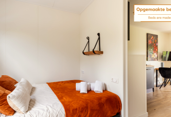 Tiny house lumineuse avec lit fait, couverture orange, coussins moelleux, serviettes et coin repas.