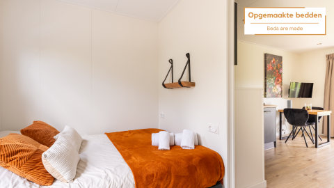 Bright tiny house with neatly made bed, orange blanket, soft pillows, towels, and small dining area.