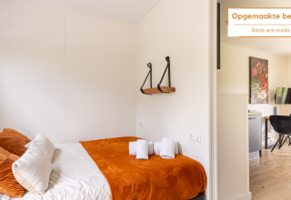 Bright tiny house with neatly made bed, orange blanket, soft pillows, towels, and small dining area.