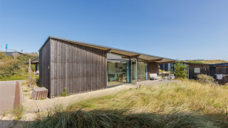 Lodge Sea House Special moderne, façade en bois et grandes baies vitrées, entouré de dunes sous ciel bleu.
