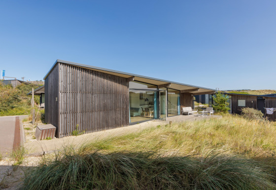 Lodge Sea House Special moderne, façade en bois et grandes baies vitrées, entouré de dunes sous ciel bleu.