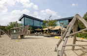 Playground with wooden climbing structures in front of a modern building at Bloemendaal aan Zee holiday park.