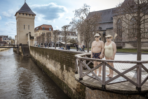 Twee realistische beelden in badpak op een balkon langs een rivier in een schilderachtig stadje.