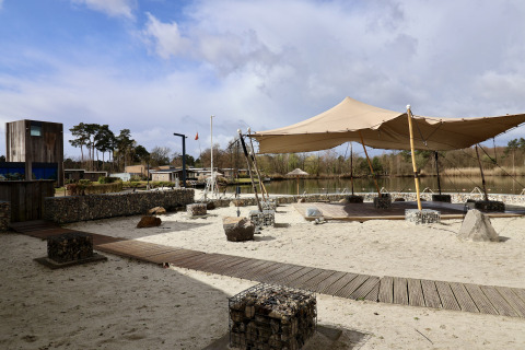 Glampinglocatie met zand, houten paden, tentoverkapping en uitzicht op een prachtig bosrijk meer.