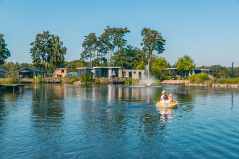 Glampingaccommodatie met moderne chalets aan een meer, twee personen varen in een opblaasboot op het water.