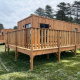 Cabaña de madera con amplia terraza en Mobi Lodge, rodeada de césped y altos árboles bajo cielo nublado.