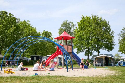 Children playing on a playground with a slide at a glamping site, surrounded by tents and green trees.