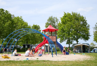 Kinder spielen auf einem Spielplatz mit Rutsche auf einem Glampingplatz, umgeben von Zelten und Bäumen.
