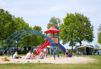 Des enfants jouent sur une aire de jeux avec toboggan dans un site de glamping entouré de tentes et d’arbres.