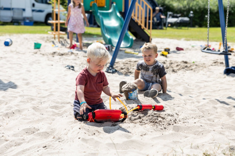 Kinder spielen im Sand auf einem Spielplatz bei einer Glamping-Unterkunft, mit Rutsche im Hintergrund.