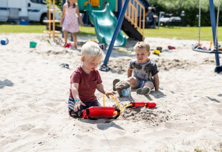 Des enfants jouent dans le sable d'une aire de jeux près d’un hébergement glamping, avec jouets et toboggan.