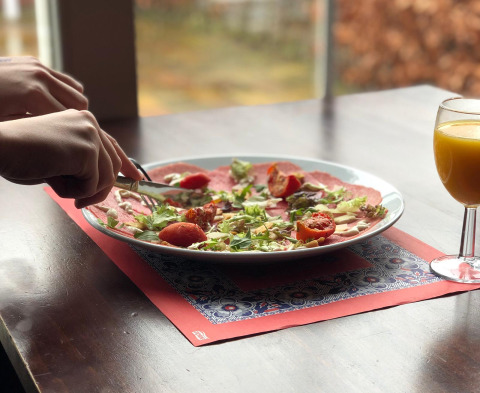 Person skærer i mad på tallerken med salat og tomater, ved siden af et glas juice i glamping-indkvartering.