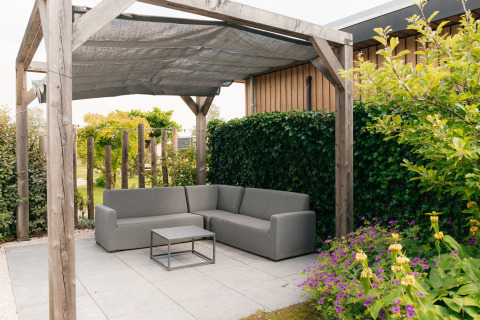 Buiten zithoek met grijze sofa onder een pergola, betegeld terras en tuin bij Veluwelodge XL in Nederland.