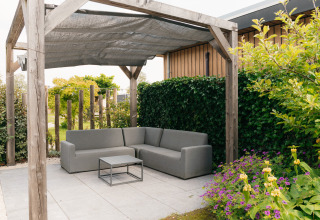 Buiten zithoek met grijze sofa onder een pergola, betegeld terras en tuin bij Veluwelodge XL in Nederland.