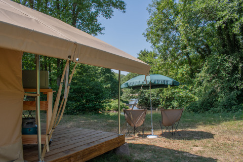 Tenda safari con terrazza in legno, sedie da campeggio e ombrellone presso Camping Le Lac de Roybon, Francia.