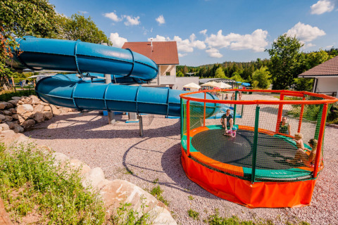 Speelterrein met grote blauwe glijbaan en ronde trampoline bij Camping La Sténiole, Grand Est, Frankrijk.