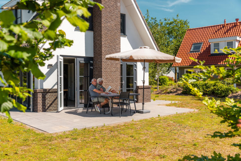 Couple âgé lisant à une table sur la terrasse d’un lodge moderne, protégés par un parasol au soleil.
