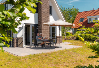 Couple âgé lisant à une table sur la terrasse d’un lodge moderne, protégés par un parasol au soleil.