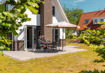 Oud echtpaar leest aan tafel op het terras van een moderne lodge met grote parasol in de zon.