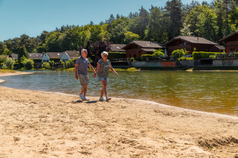 Couple de seniors marchant main dans la main sur une plage de sable devant un lac et des chalets boisés.