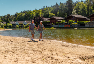 Couple de seniors marchant main dans la main sur une plage de sable devant un lac et des chalets boisés.