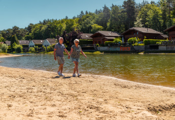 Ouder echtpaar wandelt hand in hand langs een zandstrand bij een meer met lodges en bomen erachter.