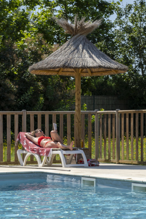Woman relaxing on a sun lounger by the pool under a thatched parasol at Seasonova Camping Le Plan d’Eau.
