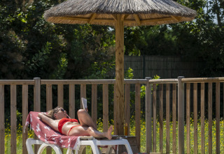 Woman relaxing on a sun lounger by the pool under a thatched parasol at Seasonova Camping Le Plan d’Eau.