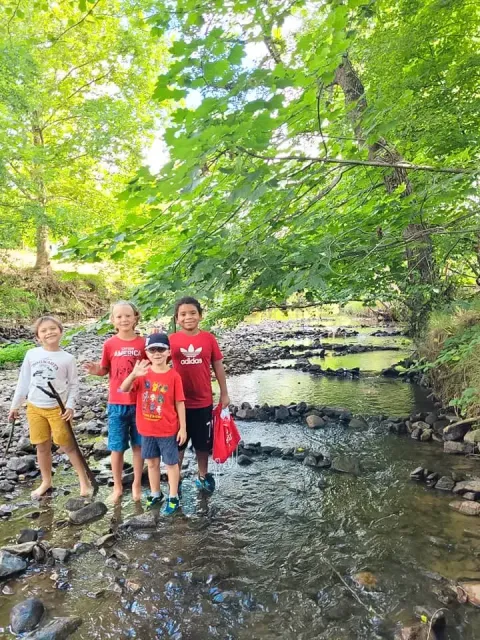 Vier Kinder spielen an einem Bach, umgeben von grünen Bäumen bei Saint-Galmier, Pays de la Loire, Frankreich.