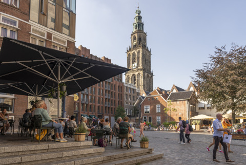 Persone sedute su una terrazza caffè vicino a una torre storica a Groningen, Paesi Bassi, in una giornata di sole.