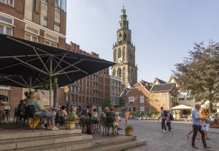 Persone sedute su una terrazza caffè vicino a una torre storica a Groningen, Paesi Bassi, in una giornata di sole.