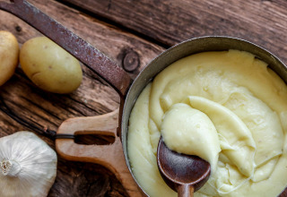 Purée de pommes de terre dans une casserole avec cuillère en bois, ail et pommes de terre sur table en bois.