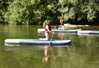 Due bambini remano in ginocchio su tavole da paddleboard al lago di Seasonova Camping Les Rives d'Olt et Aubrac in Occitania, Francia.