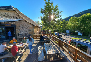 Terrasse ensoleillée avec personnes assises à Seasonova Camping Les Rives d'Olt et Aubrac, Occitanie, France.