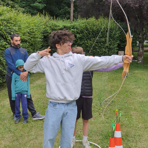 Jeune homme en sweat tire à l’arc sur une pelouse, entouré d’autres personnes lors d’un camp en plein air.