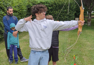 Junger Mann mit weißem Hoodie schießt mit Pfeil und Bogen auf einer grünen Wiese mit anderen Teilnehmern.
