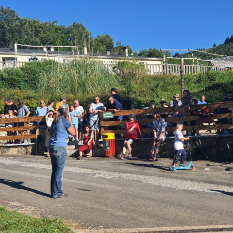 Menschen entspannen im Freien eines Ferienparks, einige sitzen, ein Junge fährt auf einem Tretroller.