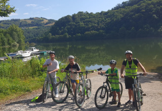 Quattro persone con biciclette e caschi vicino a un lago a Seasonova Camping Les Rives d'Olt et Aubrac in Occitania, Francia.
