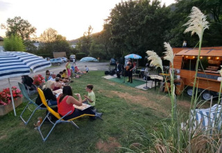 Scena all'aperto al Seasonova Camping Les Rives d'Olt et Aubrac in Occitania, Francia, con persone che mangiano e musica dal vivo.