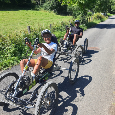 Due persone pedalano su biciclette reclinate su una strada soleggiata vicino a Seasonova Camping Les Rives d'Olt et Aubrac, Francia.