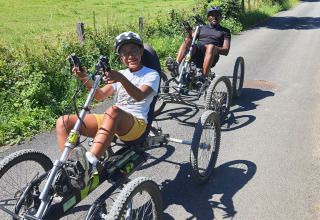 Due persone pedalano su biciclette reclinate su una strada soleggiata vicino a Seasonova Camping Les Rives d'Olt et Aubrac, Francia.