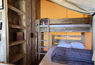 Vista interior de la tienda safari 'Slow Lodge' con litera, cama doble y estantería en Camping Seasonova.