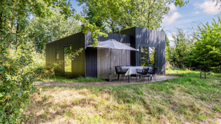 Lodge moderne Green Valley 6 noire entourée de verdure avec terrasse, table à manger et parasol.
